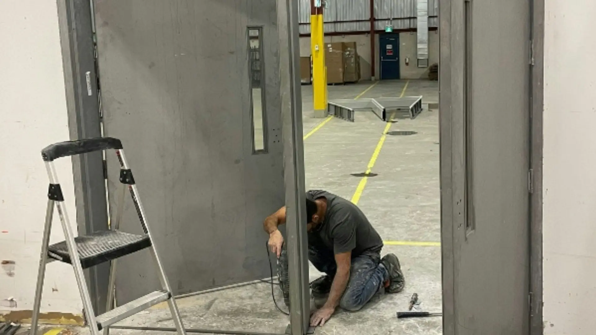 Technician kneeling to fit a commercial steel door frame, with a stepladder beside the opening