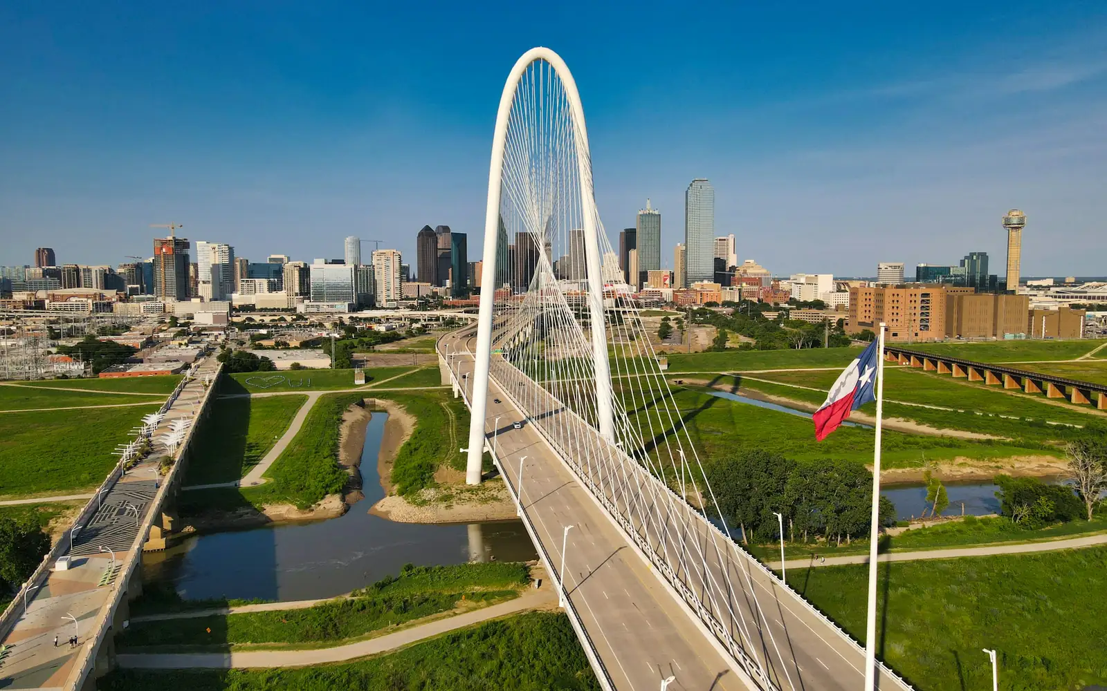 Margaret Hunt Hill Bridge and a Texas flag in front of the downtown Dallas skyline on a clear day