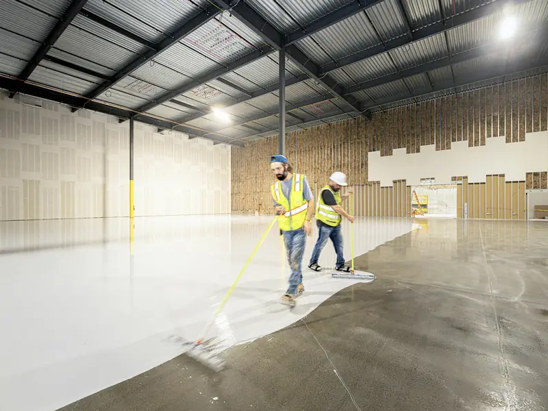 Two workers in safety vests spreading a new coating across the concrete floor of an empty warehouse